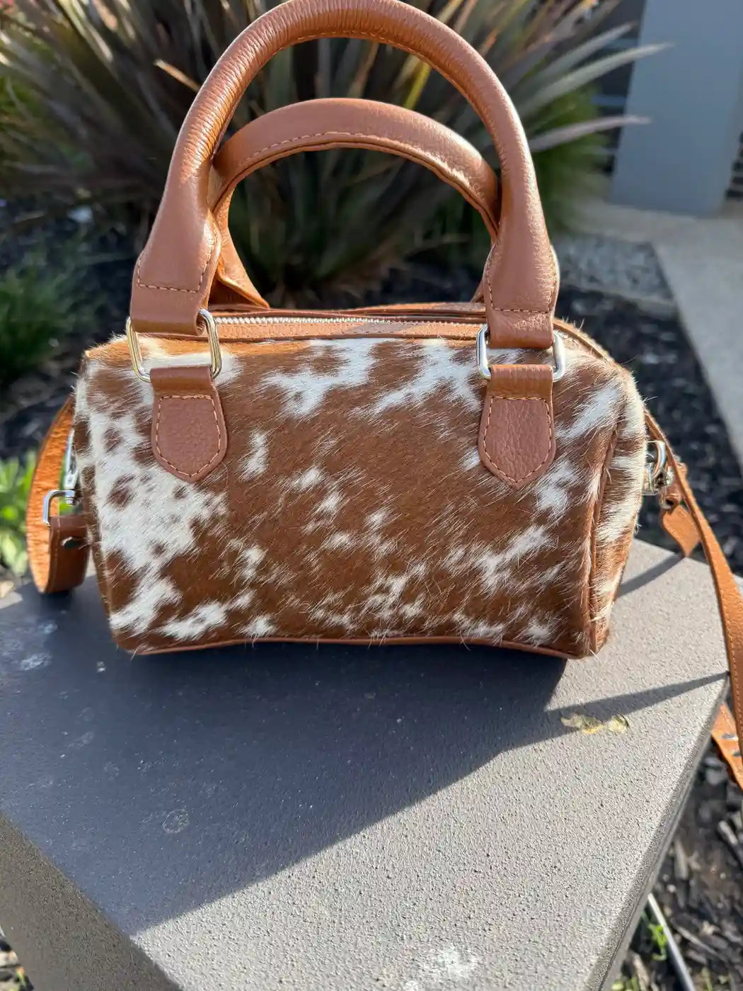 Brown and white cowhide handbag with leather handles and trim on a stone surface.