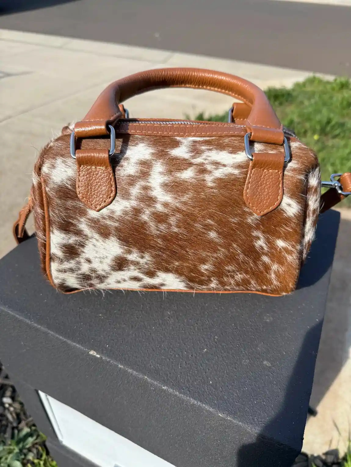 Brown and white cowhide patterned handbag on a black surface with a blurred outdoor background.