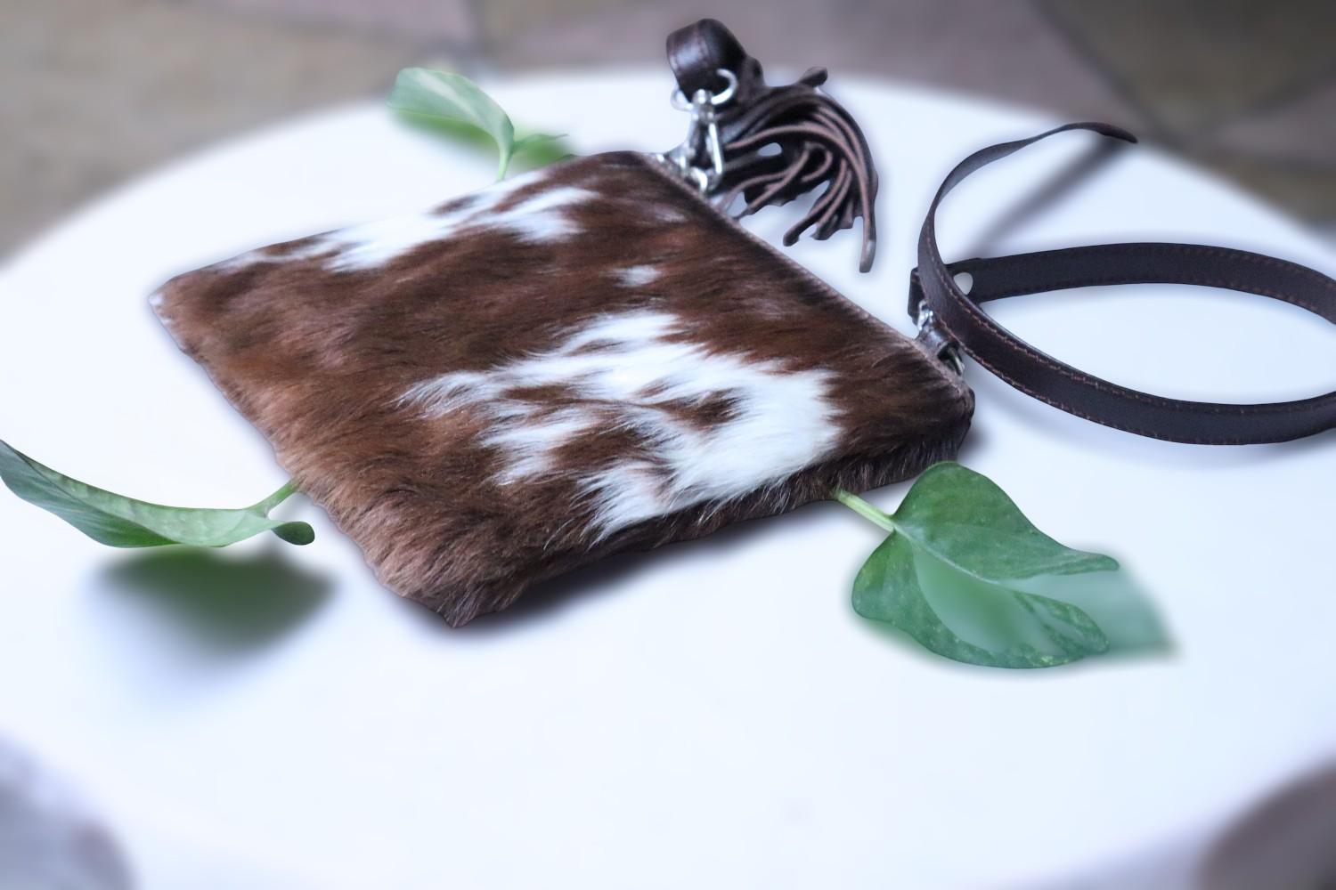 Brown and white cowhide pouch with a black strap on a white surface with green leaves.