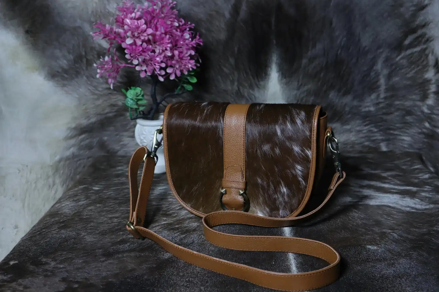 Brown cowhide leather handbag with a strap on a textured dark surface with pink flowers in the background.